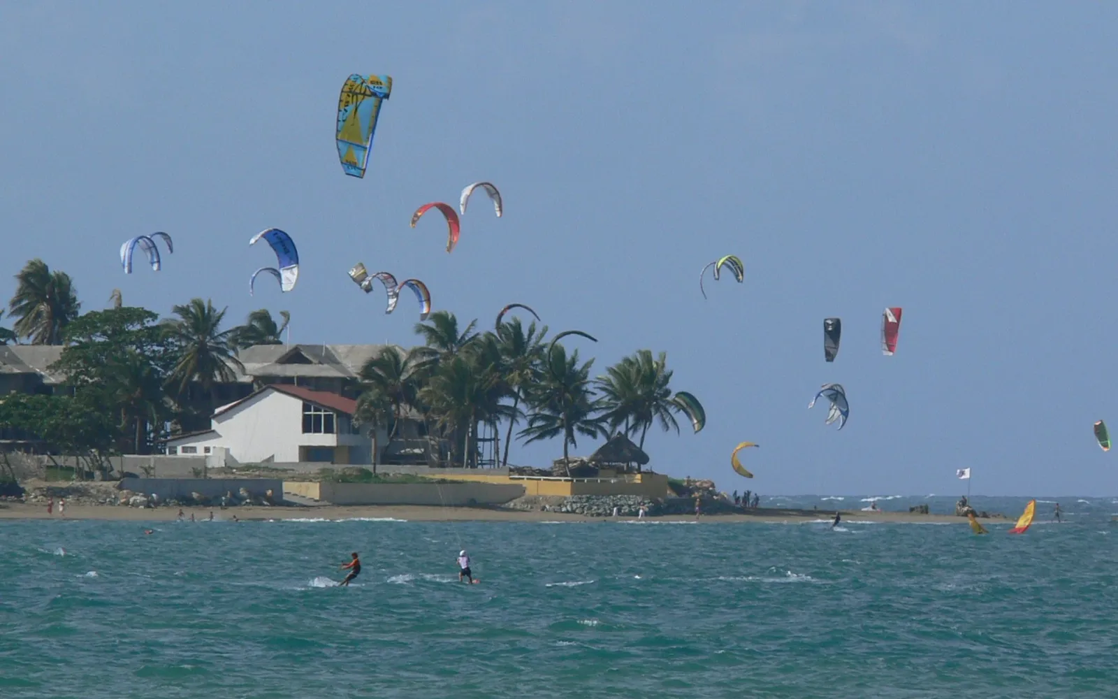 Zahlreiche bunte Kites über der Bucht von Cabarete vor palmengesäumter Küste.