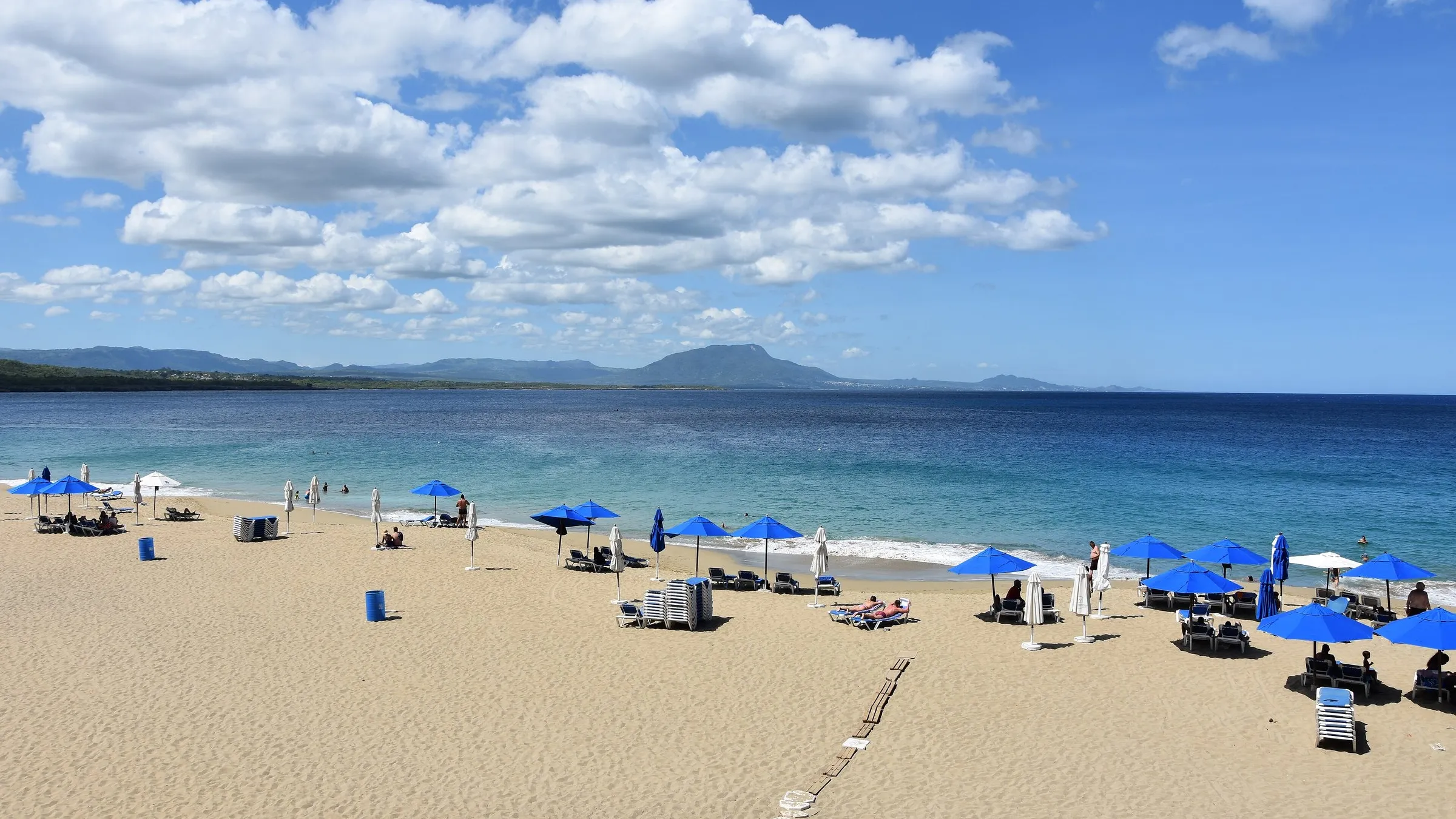 Sandstrand mit blauen Sonnenschirmen an der Bucht von Sosúa, im Hintergrund die Berge.